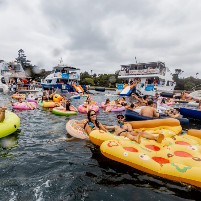 A lively scene of people enjoying a party on the water, surrounded by several boats. Many are relaxing on colorful inflatable floaties shaped like pizza, unicorns, and donuts. The sky is overcast, and the event is part of The Yacht Social Club in Sydney near a wooded shoreline.