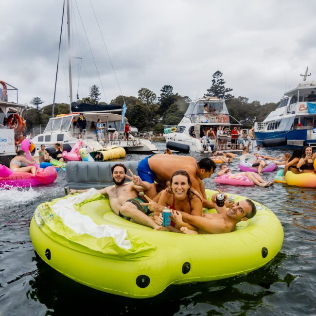 A lively scene of people enjoying a boat party on water; some are on boats, others on colorful inflatables, including a bright green avocado float. The background shows boats with festive decor and cloudy skies overhead, contributing to the festive atmosphere synonymous with Boat Parties Sydney The Yacht Social Club.