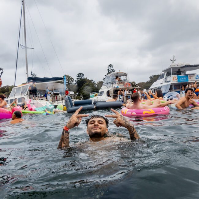 A man is waist-deep in water at a lively boat party hosted by The Yacht Social Club, with multiple colorful inflatables and several boats in the background. He is making a peace sign with both hands, surrounded by other people enjoying the event. The sky is overcast.