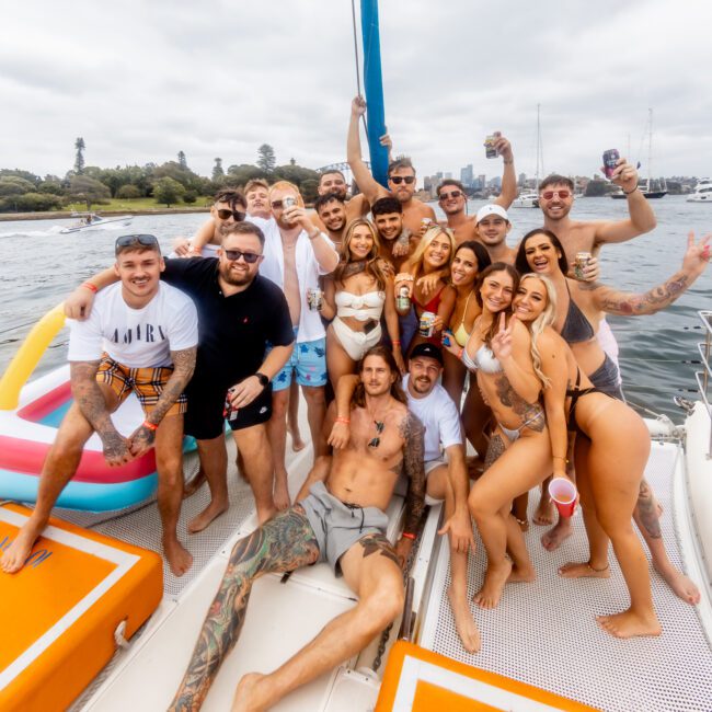 A group of people is gathered on a boat, dressed in swimsuits and casual summer outfits. They are smiling, raising drinks, and celebrating. The Yacht Social Club Event creates the perfect setting on the water with a cloudy sky overhead and a distant shoreline visible in the background.