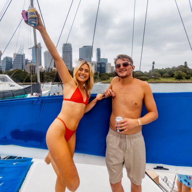 A woman in a red bikini holds up a drink and smiles, standing next to a shirtless man in sunglasses holding a can, both aboard a boat. City buildings and Sydney Harbour are visible in the background, courtesy of Luxury Yacht Rentals Sydney.