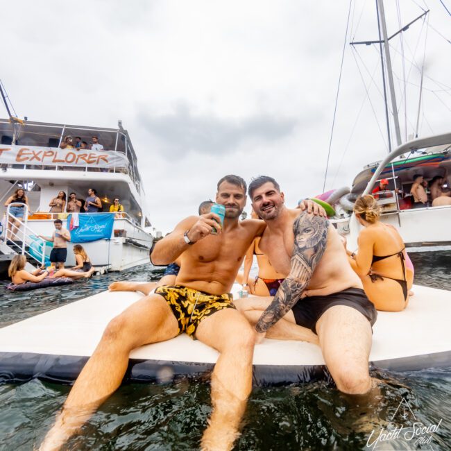 Two men sit on a floating platform in the water, smiling and posing for a photo. Other people are visible on boats around them, with one boat in the background bearing "Explorer" written on it. It appears to be a social gathering featuring The Yacht Social Club Sydney Boat Hire.