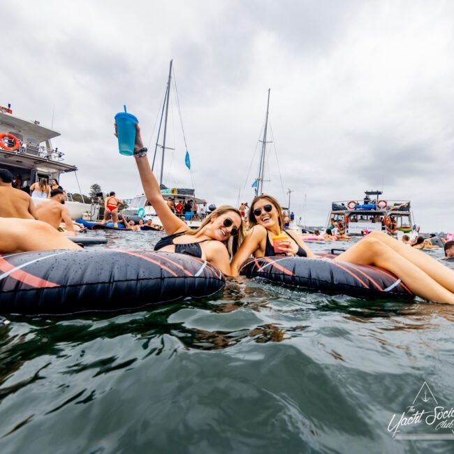 Two women in bikinis lounge on inflatable rafts, holding drinks and smiling at the camera. Behind them, a group of people are socializing on a docked boat from The Yacht Social Club. Several sailboats float in the background under a cloudy sky, making it an ideal day for Luxury Yacht Rentals Sydney.