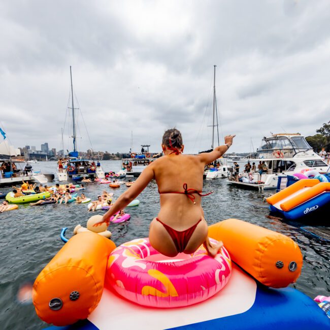 A woman in a red bikini sits on a colorful inflatable float, facing away from the camera on a crowded waterway with numerous boats and other people on inflatables. She holds up her left arm, appearing to wave or point. The sky is overcast at The Yacht Social Club Event Boat Charters.