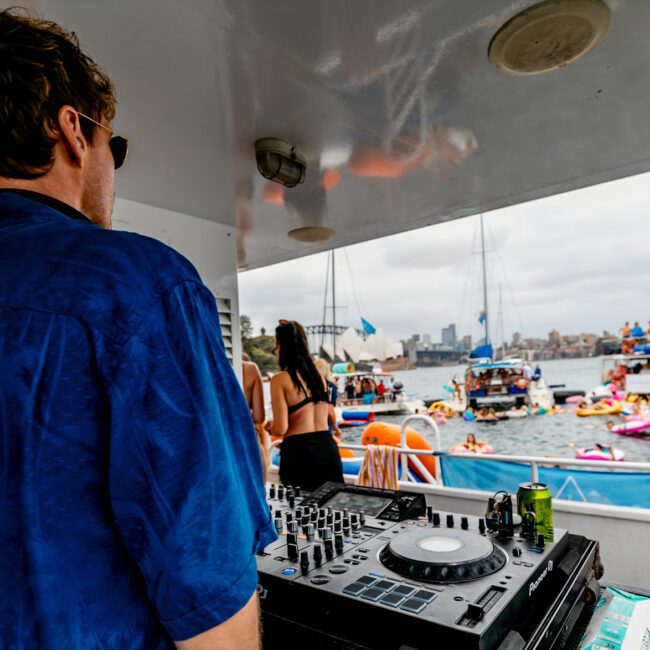 A DJ wearing a blue shirt and sunglasses is playing music on a boat at The Yacht Social Club event. In the background, numerous people on boats and inflatables are gathered in the water, enjoying the lively, festive atmosphere despite the cloudy day.