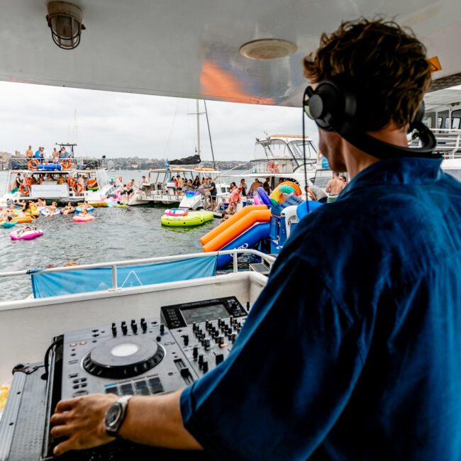 A DJ in headphones and a blue shirt stands at a mixing console on The Yacht Social Club Event Boat Charters, overlooking a lively scene of people on colorful inflatables and boats on the water. The surroundings suggest a festive atmosphere with blue and white boats nearby.