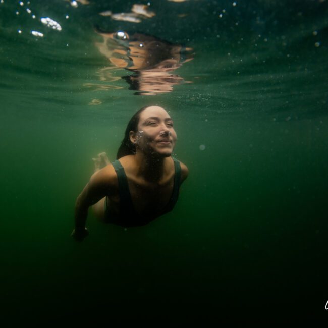 A woman is swimming underwater in a green-tinted environment, her face turned upwards and smiling. She is wearing a dark swimsuit. The logo in the bottom right corner reads: "Sydney Harbour Boat Hire The Yacht Social Club".