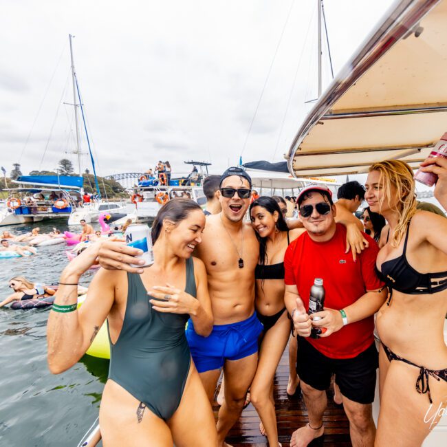 A group of people enjoying a party on a boat rented from The Yacht Social Club. They are dressed in swimwear, holding drinks, and smiling at the camera. The background shows other boats and people swimming or floating on inflatable rings in the water. The sky is cloudy.