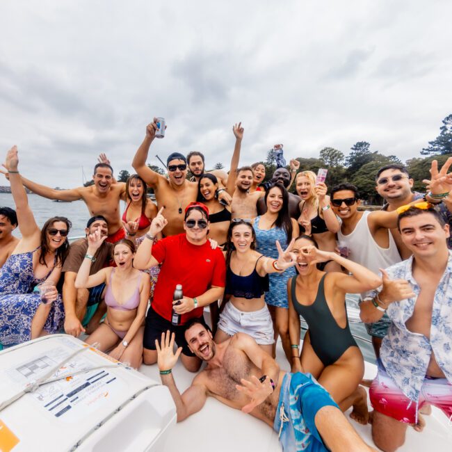 A group of people are enjoying a boat party by the water, courtesy of Sydney Harbour Boat Hire The Yacht Social Club. They are smiling, laughing, and making celebratory gestures. Some are holding drinks, while others pose energetically in their swimwear against a backdrop of cloudy skies and distant trees.