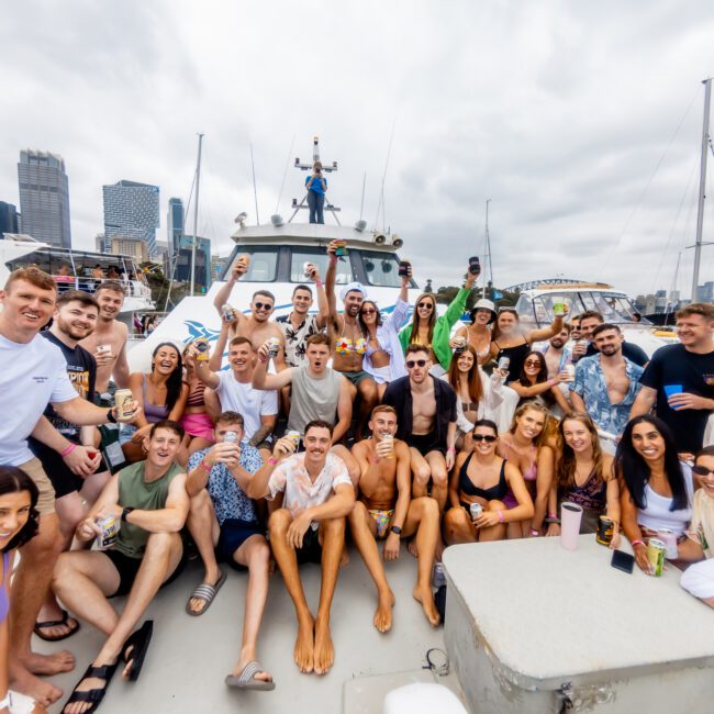 A large group of people are gathered on a boat, smiling and posing for the camera. The background features a city skyline and other boats on the water. The atmosphere is lively, with everyone appearing to enjoy a social event or celebration hosted by Boat Rental and Parties Sydney.