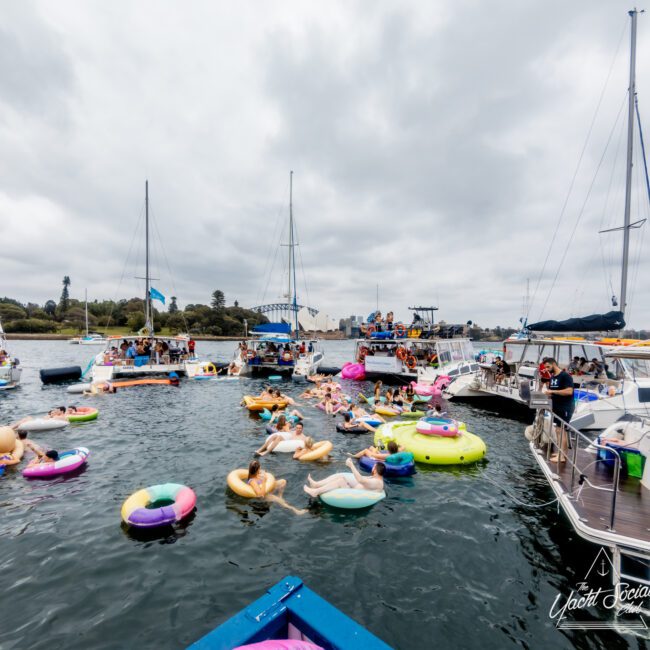 A group of people enjoy a social gathering on a lake, floating on various colorful inflatables. Several boats and yachts from Sydney Harbour Boat Hire The Yacht Social Club are anchored nearby. The sky is cloudy, and there are trees in the background.