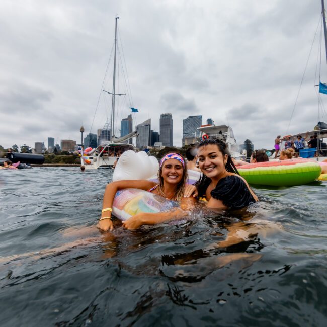 Two smiling women enjoy a water party, hugging each other while floating on inflatable mattresses. Dozens of other people are seen on the water or nearby boats. The city skyline with high-rise buildings is visible in the background under a cloudy sky, showcasing the vibrant atmosphere of Boat Parties Sydney The Yacht Social Club.