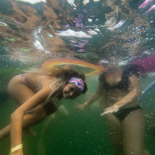 Underwater shot of one woman in a colorful bikini and headband swimming toward the camera while another woman in a black swimsuit hovers in the background. The scene is lively with light reflections from the water's surface above. The Yacht Social Club Sydney Boat Hire logo is visible.