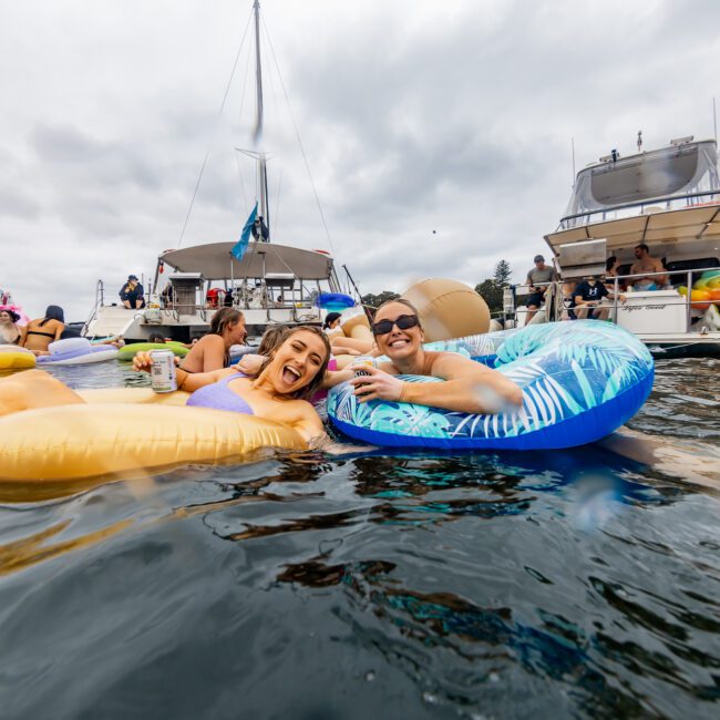 Two people relax on inflatable floats in the water, smiling and holding drinks. Surrounding them are several boats, and others are swimming or on floats. The sky is overcast, and the atmosphere is lively and festive. A logo reads "The Yacht Social Club," known for Luxury Yacht Rentals Sydney.