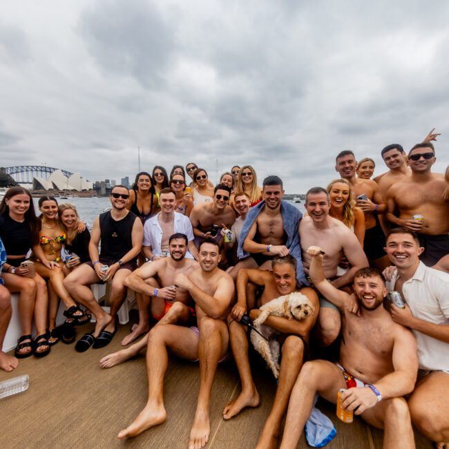 A large group of people, wearing swimwear, are gathered on a boat from The Yacht Social Club, smiling and posing for a photo. The sky is overcast, and a bridge and waterside buildings are visible in the background. Some people are holding drinks and one person is holding a dog.