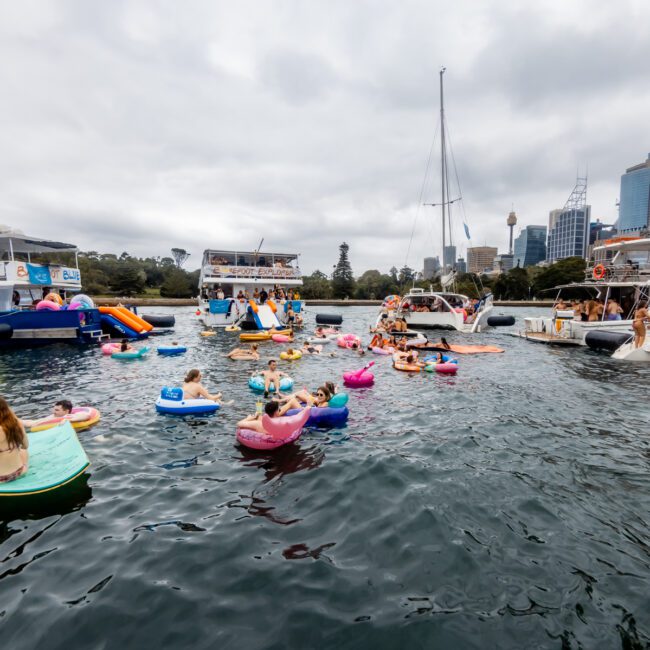 A lively scene of a party on the water with people on various inflatable rafts and floats, surrounded by multiple anchored boats. The background features city skyscrapers under a cloudy sky. The event, organized by The Yacht Social Club Sydney Boat Hire, appears joyful with attendees enjoying themselves.