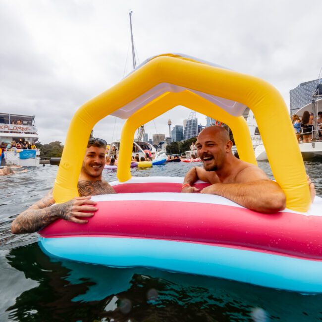 Two men are smiling and relaxing in a small inflatable raft shaped like a house while floating on water. There are other people enjoying the water nearby, and boats from Luxury Yacht Rentals Sydney are visible in the back. The sky is overcast, adding a calm ambiance.