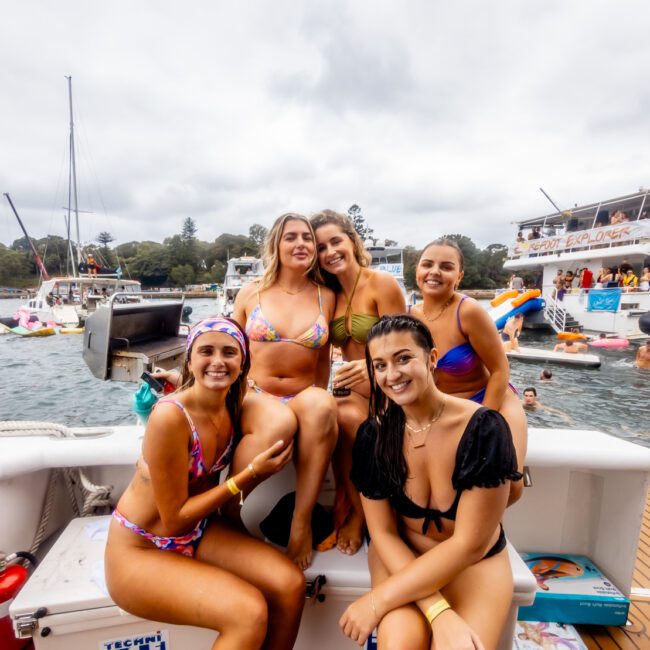 A group of five young women in swimsuits sit and stand on a boat, smiling at the camera. Multiple other boats with people on them are visible in the background, creating a festive, lively atmosphere. It appears to be a party or gathering by The Yacht Social Club Sydney Boat Hire on the water.