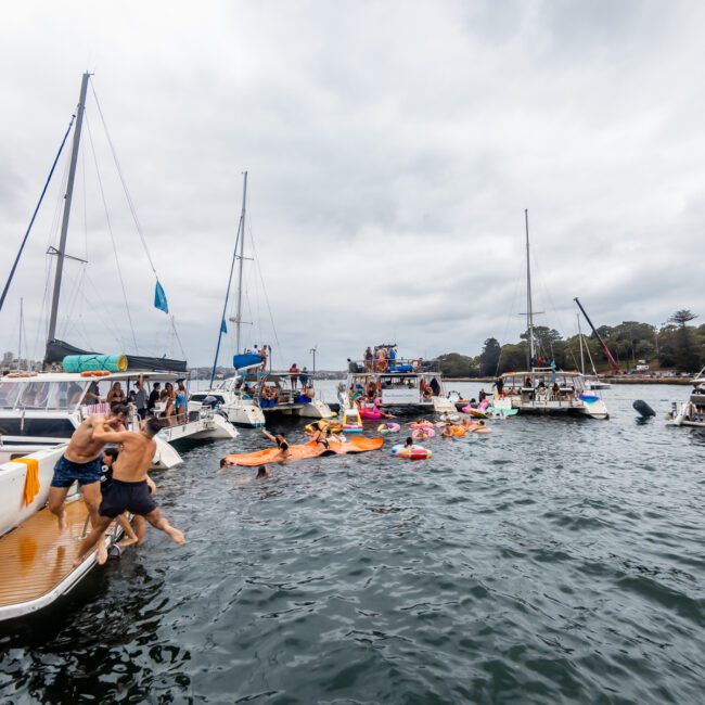 A lively scene of people enjoying a day on the water with several boats and yachts anchored close together. Many are swimming, using floaties, and socializing in the water. The sky is cloudy, and the image promotes a sense of fun and community, perfect for Sydney Harbour Boat Hire The Yacht Social Club events.