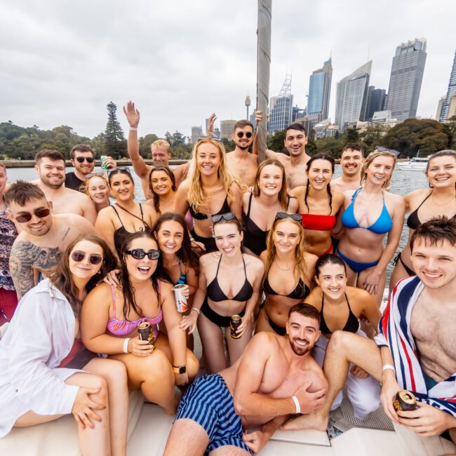 A large group of people in swimsuits are gathered and smiling on a boat in a harbor. The backdrop features a city skyline with several tall buildings and green areas near the water. Some people hold drinks, and there's a festive atmosphere, typical of Sydney Harbour Boat Hire The Yacht Social Club events.