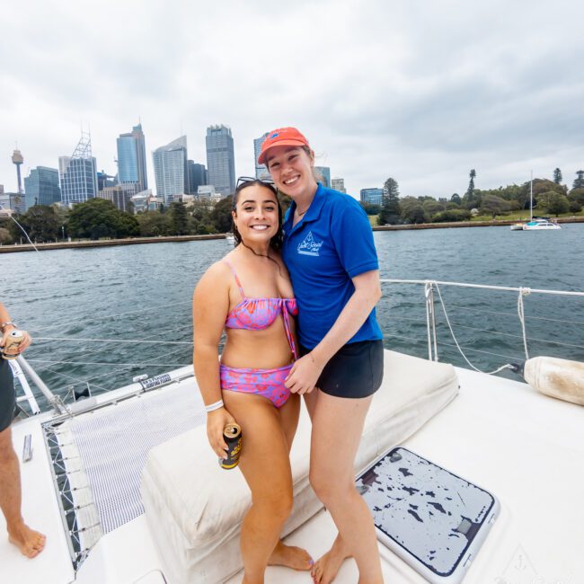 Two people on a boat with a cityscape in the background. One person is wearing a pink swimsuit and holding a beverage, while the other is dressed in a blue polo and black shorts. They are standing close together, smiling, with water and buildings visible behind them—an ideal scene for The Yacht Social Club Event Boat Charters in Sydney.