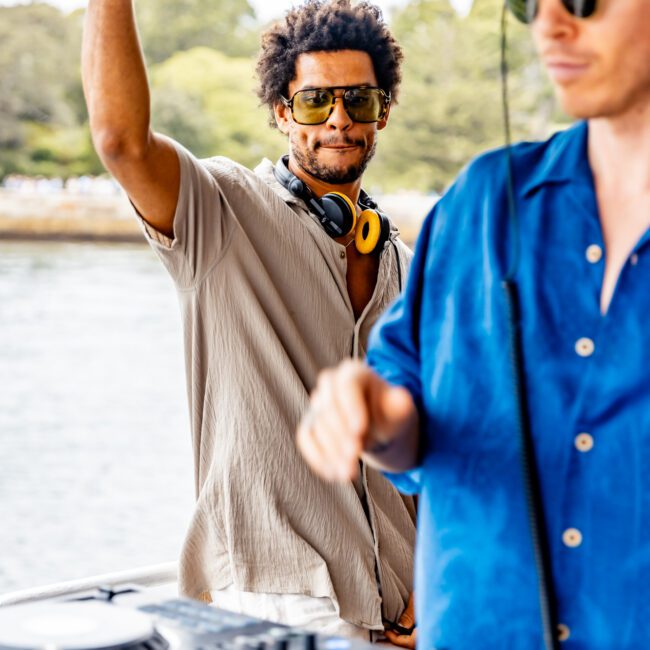 Two DJs are performing on a boat with water and greenery in the background. One DJ, wearing a beige shirt and yellow sunglasses, raises his arm, while the other, in a blue shirt and sunglasses, is focused on the DJ console. "The Yacht Social Club" is visible in the lower right corner at their Sydney Harbour Boat Hire event.