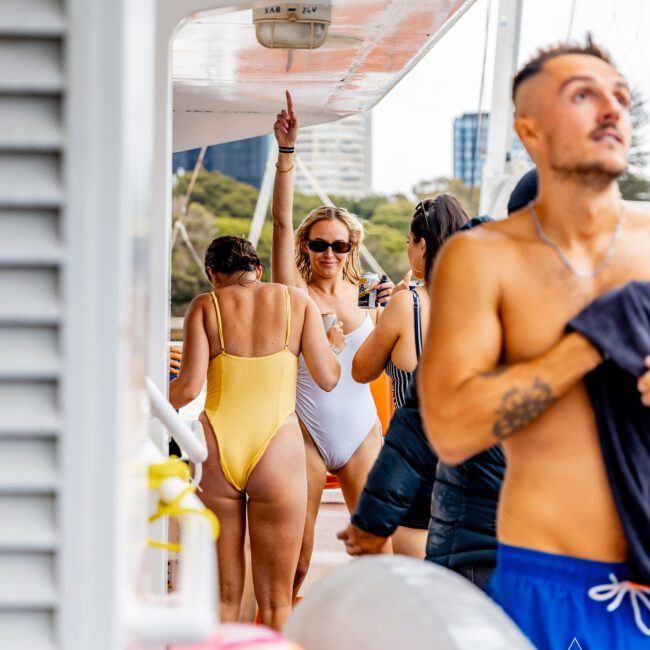 Group of people on a boat deck, enjoying a sunny day. A woman in a white swimsuit holds a drink and gestures upwards. Another woman in a yellow swimsuit stands nearby, while a man in blue shorts holds a towel. Inflatable toys are visible in the foreground at The Yacht Social Club Event Boat Charters.