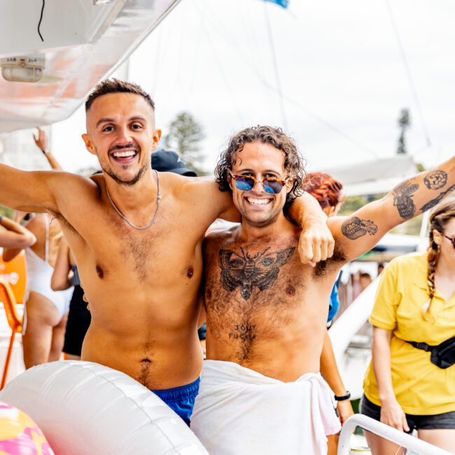 Two shirtless men, one with a towel around his waist and sunglasses, stand smiling with arms around each other on a boat. The scene includes other people, a partial view of an inflatable float, and maritime elements in the background, capturing the essence of The Yacht Social Club Sydney Boat Hire experience.