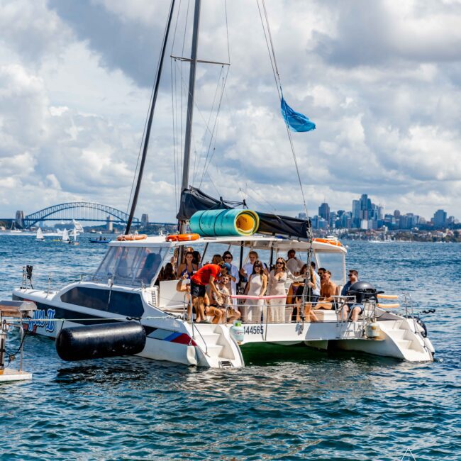 A catamaran with several people on board is sailing in a body of water. City buildings and a prominent bridge are visible in the background under a partly cloudy sky. A blue flag flies from the boat's mast. The calm water reflects the clear sky, making it perfect for The Yacht Social Club Sydney Boat Hire adventure.