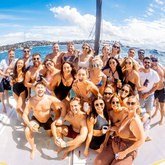 A group of around 25 smiling people is gathered on a boat, enjoying a sunny day. They are posing for the photo, with a mix of men and women dressed in swimwear. The background shows blue waters and distant buildings under a clear, bright sky. It's an event by The Yacht Social Club Sydney Boat Hire.