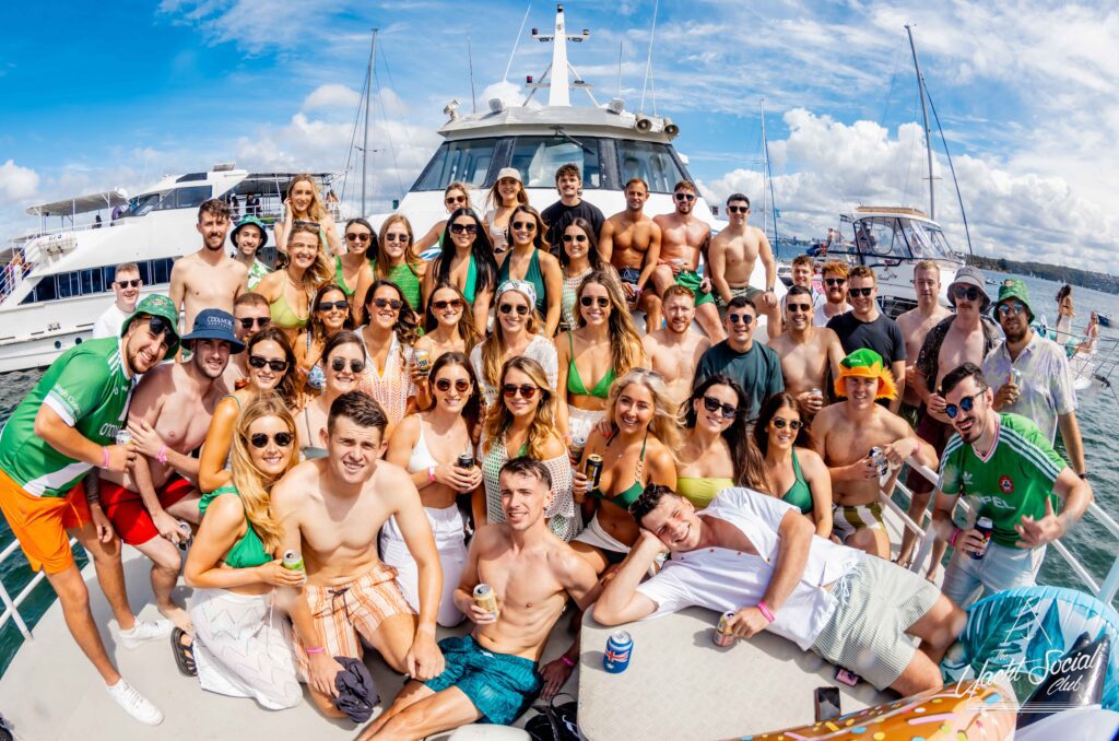 A large group of people wearing sunglasses and summer clothing are gathered on the deck of a boat, smiling and posing for a photo during The Yacht Social Club event. The sky is blue with some clouds, and the water is visible in the background. Some are holding drinks, and one person has a camera.