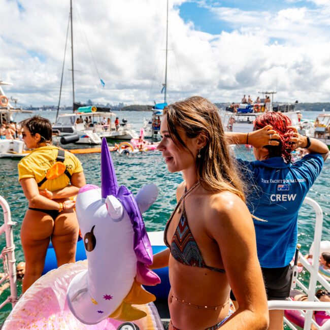 A woman in a bikini stands on a boat, holding an inflatable unicorn pool float and a beverage can. Other people and boats in the background are enjoying the sunny day on the water. A crew member in a blue shirt is visible alongside sailboats and a partly cloudy sky, showcasing Luxury Yacht Rentals Sydney.