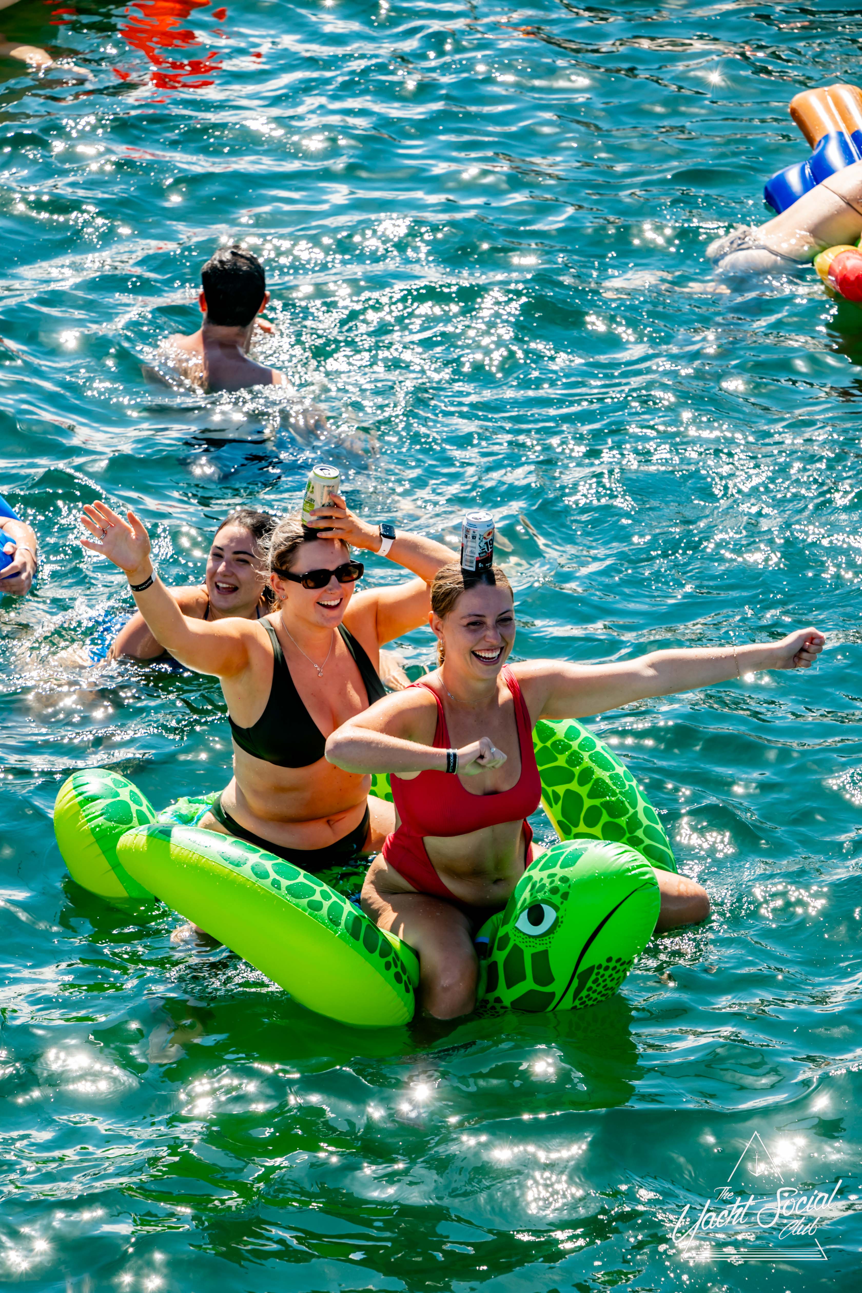 Two women in swimsuits laugh and pose on an inflatable green turtle in a sunlit body of water. One woman balances a drink can on her head while the other flexes her arm. Other swimmers and inflatable toys are visible in the background, giving it a relaxed vibe reminiscent of The Yacht Social Club Event Boat Charters.