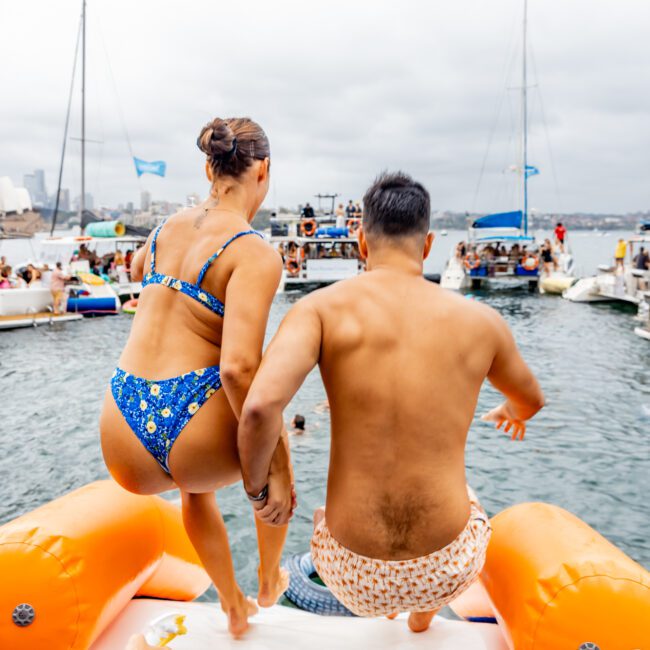 Two people in swimsuits are about to jump off an inflatable platform into a body of water during The Yacht Social Club Event Boat Charters. Boats and other people are visible in the background on this overcast day.