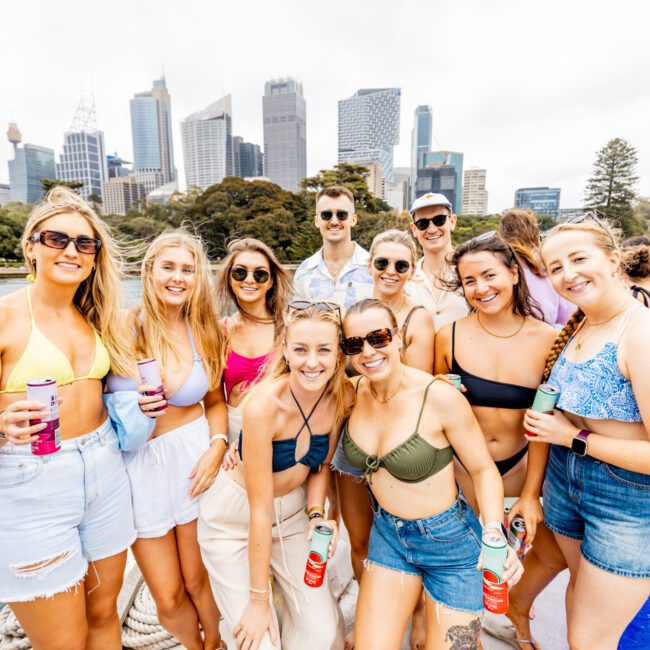 A group of friends pose happily on a boat with drinks in hand, dressed in casual summer attire. With a city skyline and waterfront park as the backdrop, the atmosphere at The Yacht Social Club Event Boat Charters is festive and joyful.
