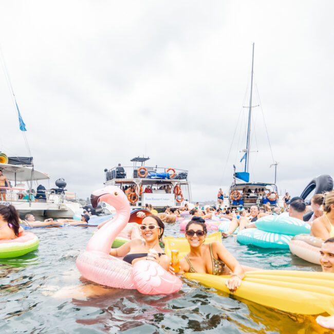 A lively scene at The Yacht Social Club event features many people enjoying the water on various inflatable floats, including a large pink flamingo. The sky is overcast. Several boats are anchored in the background, and guests are socializing and having fun.