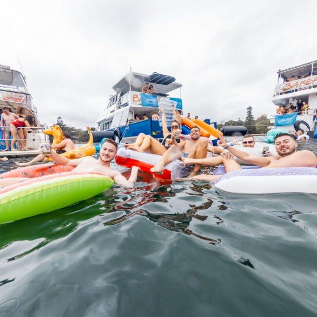 A group of people floats on inflatable loungers in a body of water near several docked boats on a cloudy day. They are smiling, holding drinks, and appear to be enjoying a social gathering hosted by The Yacht Social Club. The scene is lively and festive.