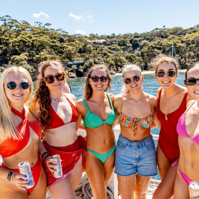 A group of six smiling women in swimsuits pose together on a boat, enjoying a sunny day on the water with a lush, green shoreline and trees in the background. They are wearing sunglasses, some holding canned beverages—a perfect day at The Yacht Social Club Event Boat Charters.