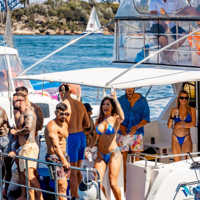 A group of people are enjoying a sunny day on a boat from The Yacht Social Club Sydney Boat Hire, dressed in swimwear. Some are standing on the deck, while others are sitting or holding drinks. A bridge and trees are visible in the background. The atmosphere appears lively and festive.