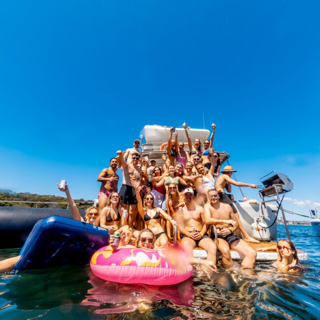 A large group of people, adults and children, are gathered on a boat and in the water, enjoying a sunny day. Some are holding drinks, and a person floats on a pink inflatable in the water. The clear blue sky adds to the joyful atmosphere, showcasing Boat Parties Sydney with The Yacht Social Club.