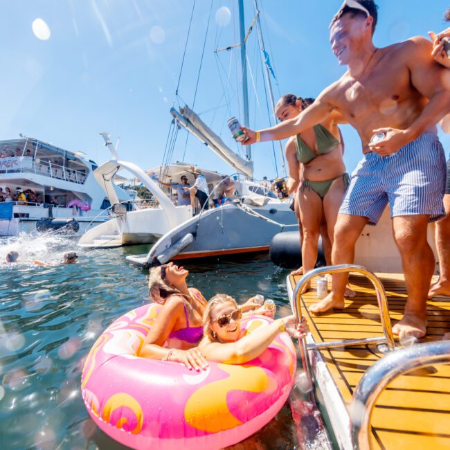 A group enjoys a sunny day on a docked boat at The Yacht Social Club Sydney Boat Hire. Two women in swimsuits relax in a pink inflatable ring in the water, smiling as a man in patterned swim trunks extends a drink toward them. Other boats and people can be seen in the background.