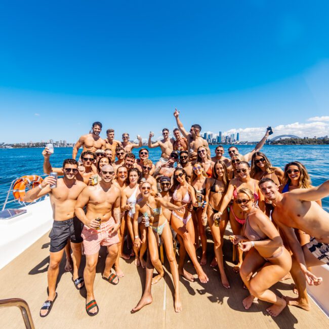 A large group of people in swimwear are gathered on a boat under a clear blue sky. They are smiling, holding drinks, and posing for the camera. A city skyline and a bridge are visible in the background over the water. It appears to be a sunny day at The Yacht Social Club Sydney Boat Hire event.