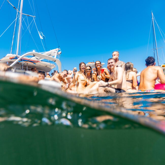 A group of people enjoying a sunny day in the water beside boats. The image captures people swimming and gathering close together, smiling and relaxed, with clear blue skies overhead and parts of sailboat masts visible in the background, showcasing a lively scene at The Yacht Social Club Event Boat Charters.