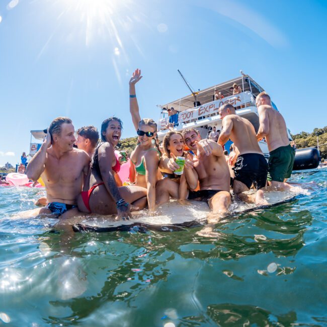 A group of people in swimsuits enjoying a sunny day on the water, sitting and standing on an inflated device. They are laughing, posing, and holding drinks, with a boat labeled "EXPLO..." nearby. The scenic forested shoreline adds to the festive atmosphere of Sydney Harbour Boat Hire The Yacht Social Club.