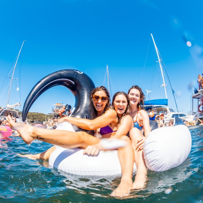 Three women in swimwear laugh and smile while sitting on an inflatable swan float in the ocean. Boats and other people swimming are visible in the background against a clear blue sky, typical of a day with The Yacht Social Club's Sydney Harbour Boat Hire.