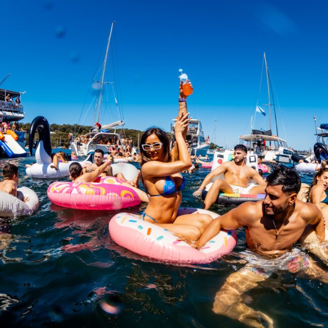 A lively scene of people enjoying a summer boat party. They are in the water, floating on inflatable rings of various shapes and colors, with boats anchored in the background. The sky is clear and bright blue, and everyone appears to be having fun at a The Yacht Social Club Event Boat Charters gathering.