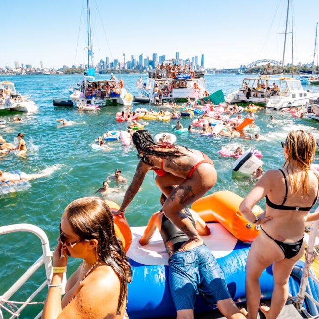 A lively scene on a sunny day with numerous people enjoying a boat party on a river. Several individuals are on inflatables in the water, while others are on boats and a floating platform. In the background, Sydney Harbour Boat Hire The Yacht Social Club events enliven the city skyline under a clear blue sky.