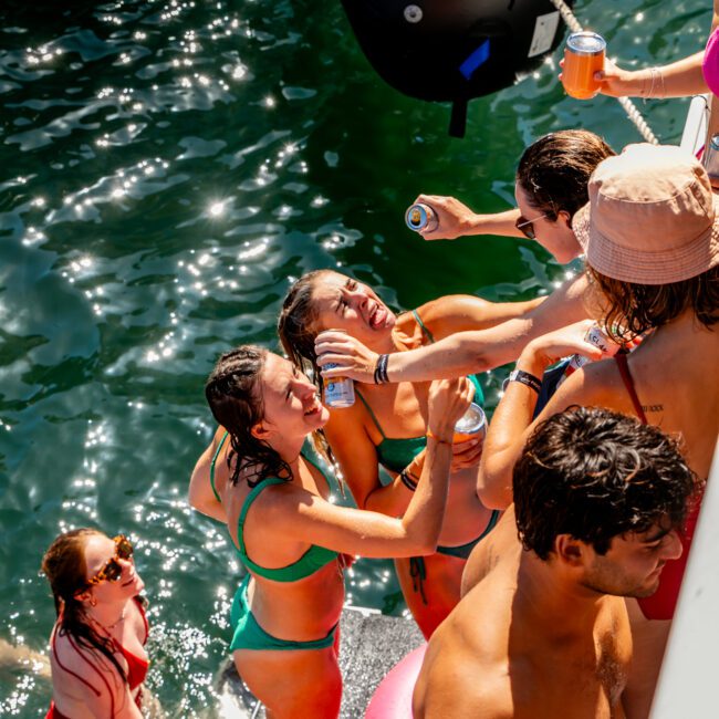 A group of young adults is enjoying a sunny day on the water, swimming and holding drinks. Some are in the water, while others are on a boat from The Yacht Social Club. They are all in swimwear and appear to be having a good time under the bright sun.