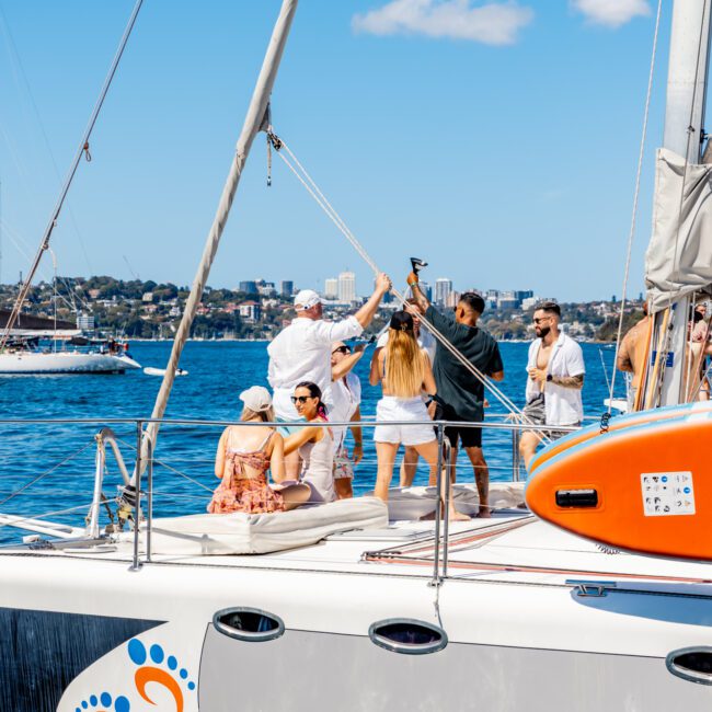 A group of people is gathered on a sailboat under a clear blue sky, adjusting the sail and socializing. The boat glides on calm water with other boats and a cityscape visible in the background. Featuring a logo with an orange and blue footprint, it's perfect for Boat Parties Sydney The Yacht Social Club events.