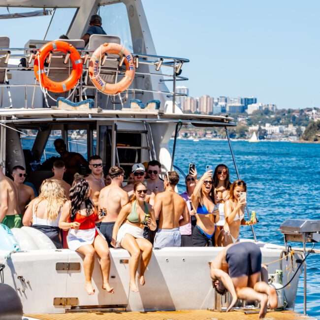 A group of people enjoying a sunny day on a boat from The Yacht Social Club, with some lounging and talking while others are preparing to swim. One person is diving off the back into the blue water. The boat has life rings, and a cityscape can be seen in the background.